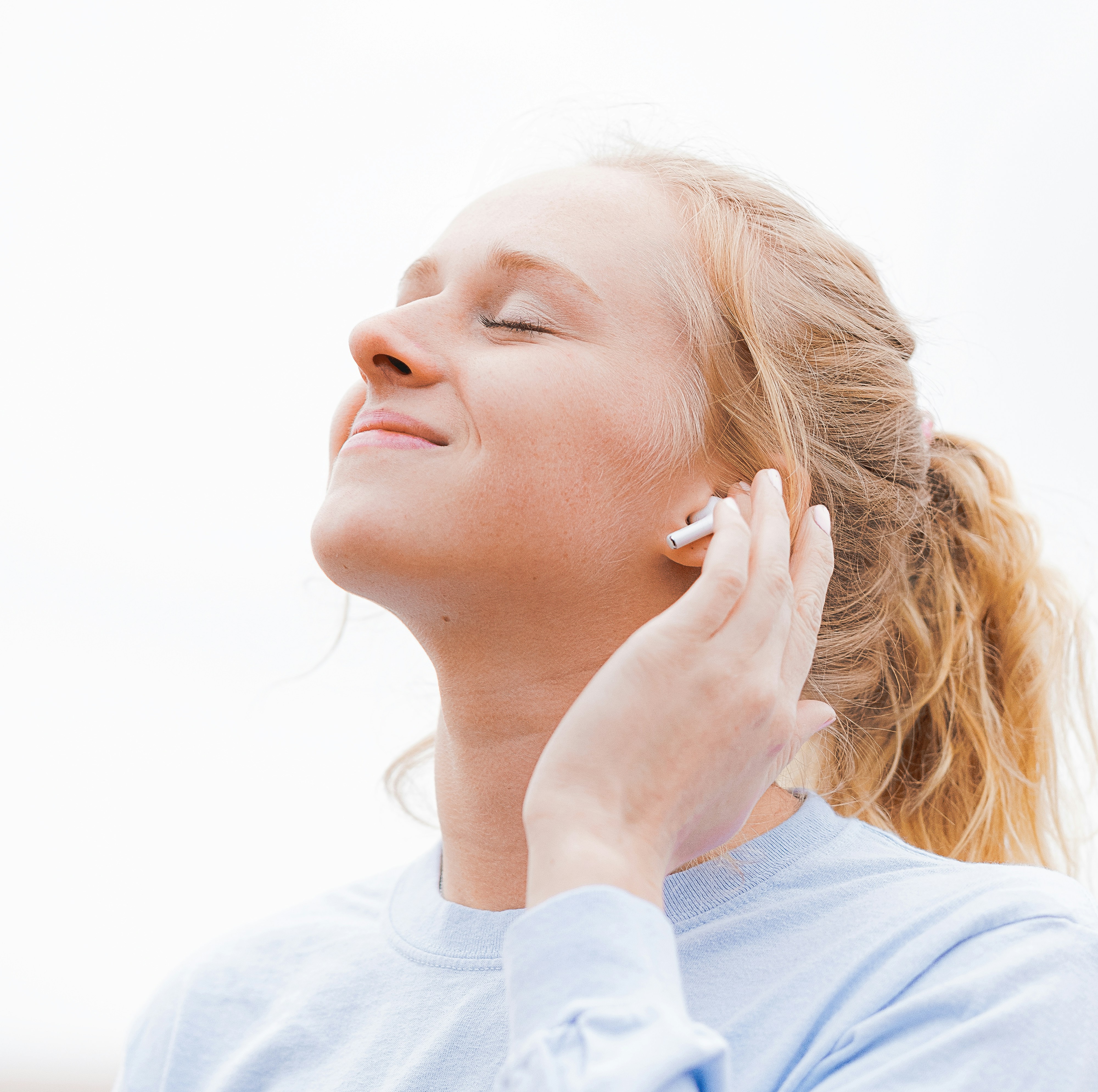 A white woman with her hair in a pony tail smiles with her eyes closed as she touches an airpod in her left ear.