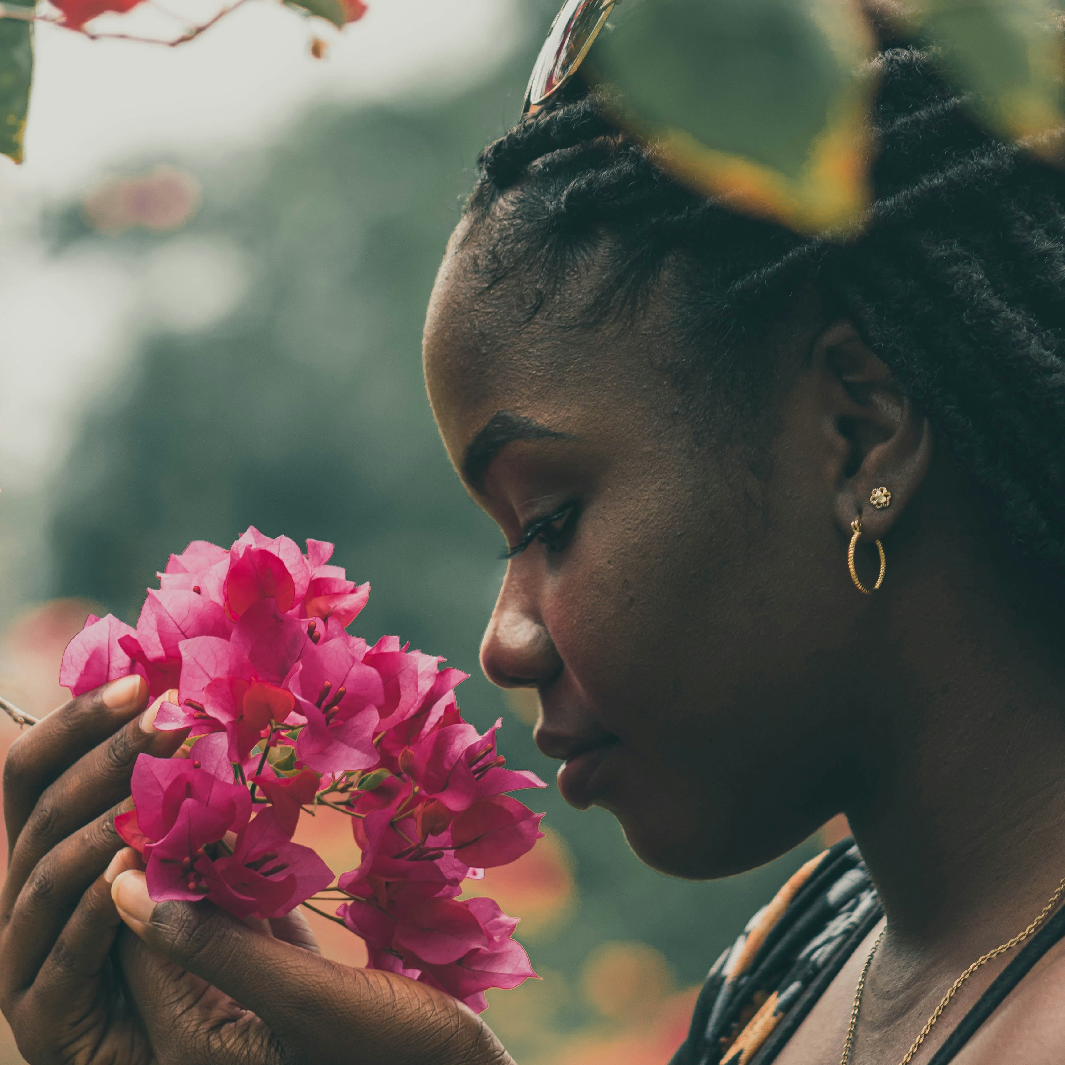 A black woman in a sundress holds her nose up to a bunch of hot pink bougainvillea blossoms.