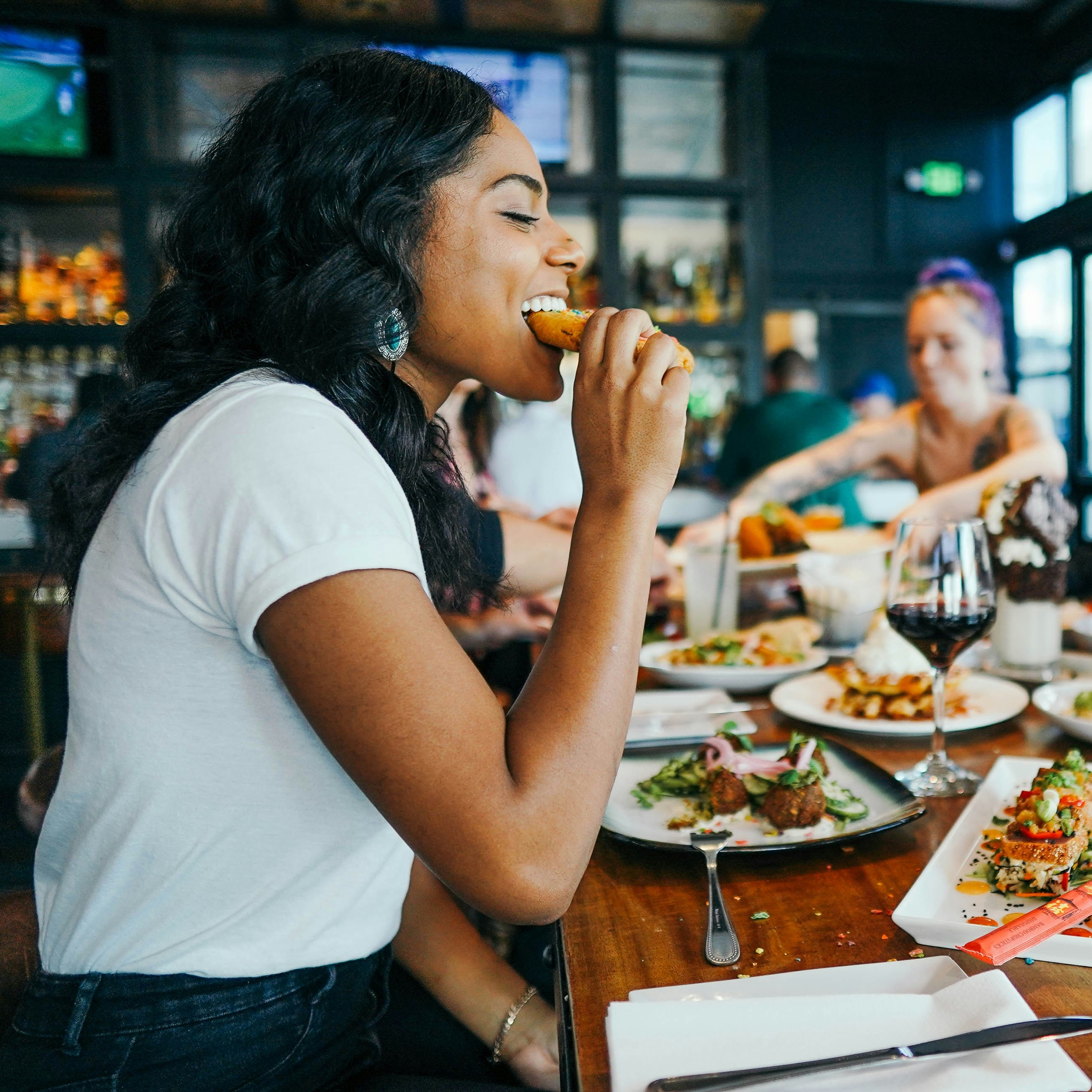 A woman in a white t-shirt with warm brown skin and long dark hair takes a bite out of a piece of handheld food at a busy gastropub.