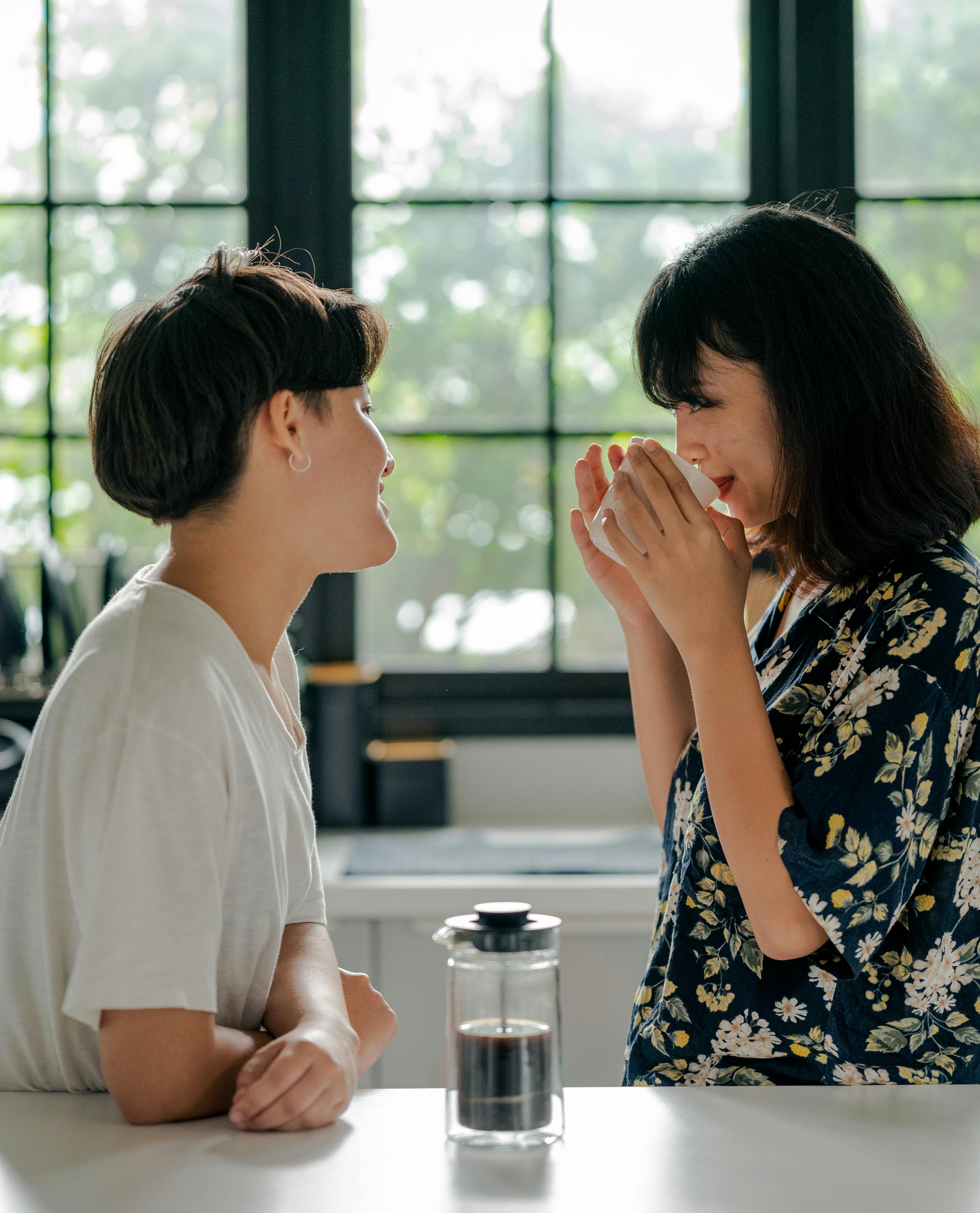 Two asian women stand in an expensive kitchen with a press pot of coffee on the counter. One of them drinks coffee while they both smile.