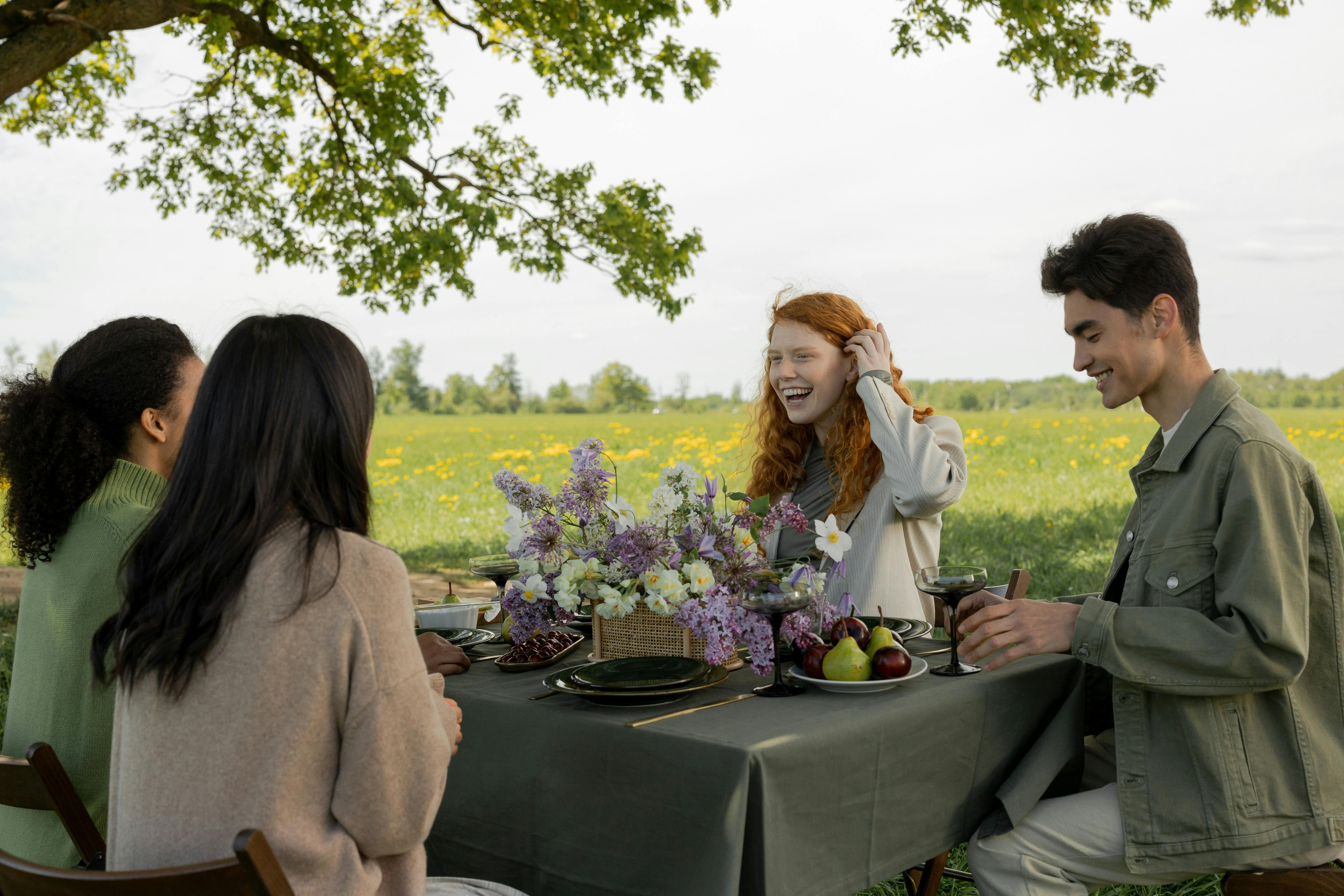 Four young people sit around a table under a tree in the springtime. On one side, a pale woman with red hair is laughing beside a smiling young man with dark hair and a green jacket. On the other side, a we see the backs of two more young women with dark hair wearing sweaters.The table has a table cloth and is set with lots of spring flowers.