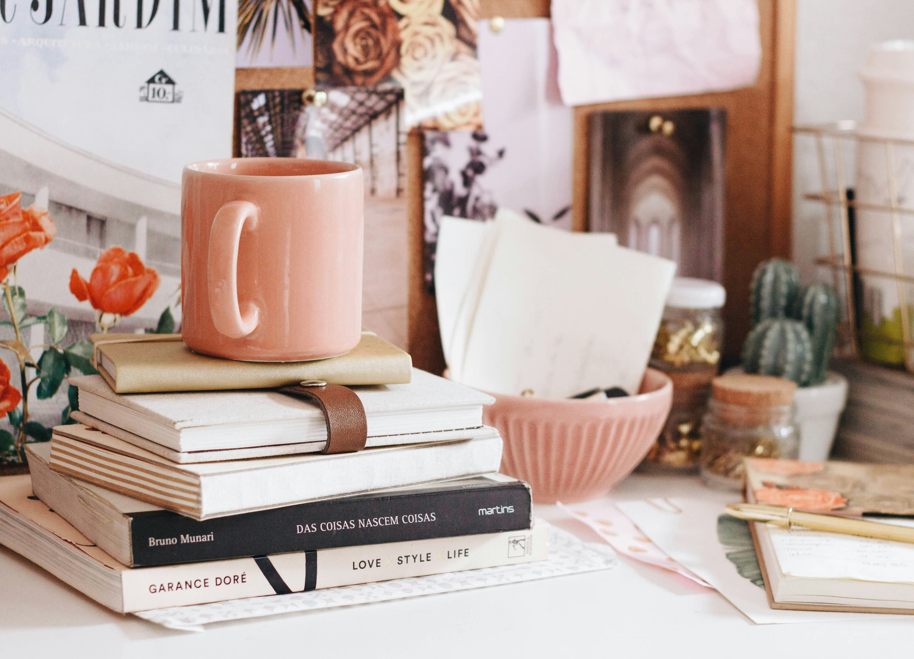 A photo of someone's workspace with a lot of brown, white, and warm pink. We see a pink coffee mug on top of a stack of books, a pink bowl full of letters, a small cactus, several jars, an open notebook, some loose papers, and, in the back, a bulletin board leaned up against the wall with several photos of architecture and flowers pinned to it.