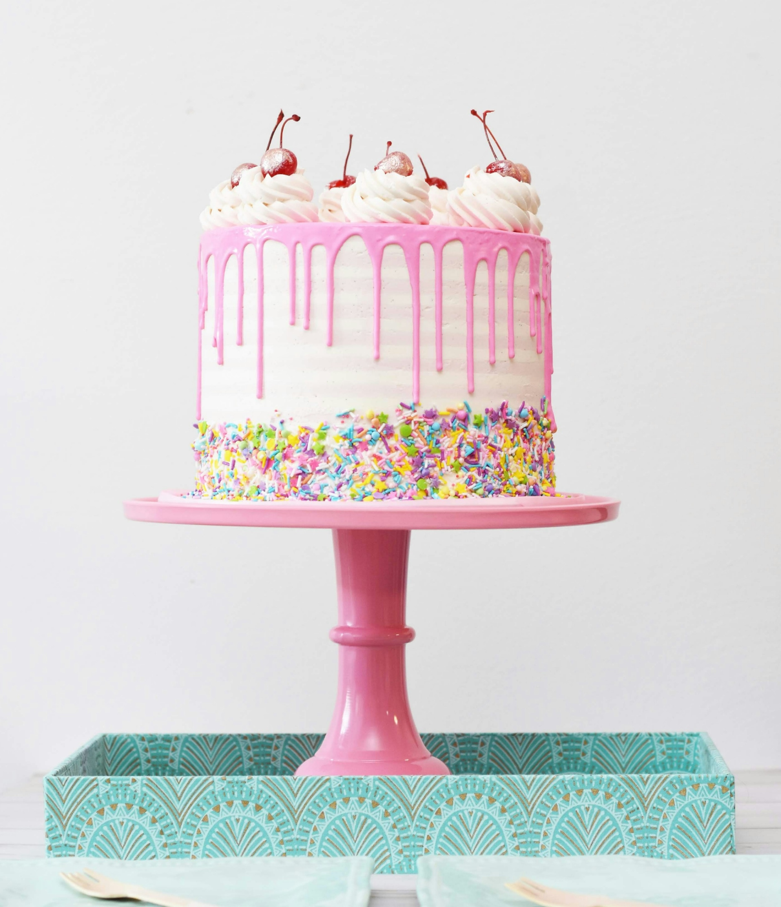 A fancy cake on a pink pedestal. It has pink and white icing and rainbow sprinkles. On top, there are dollops of whipped cream and several cherries.