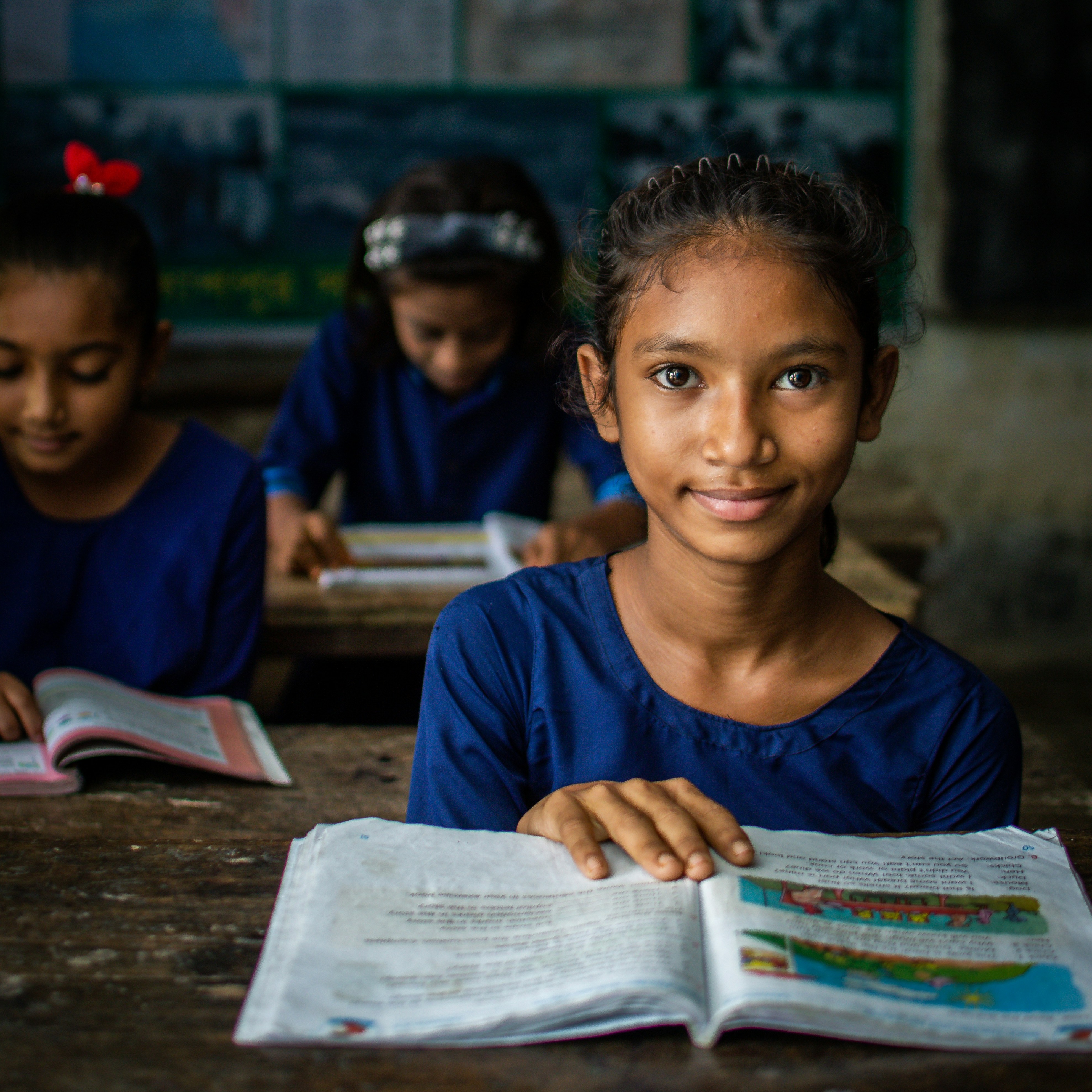 A photo of a smiling girl with brown skin sitting at a desk with a schoolbook.